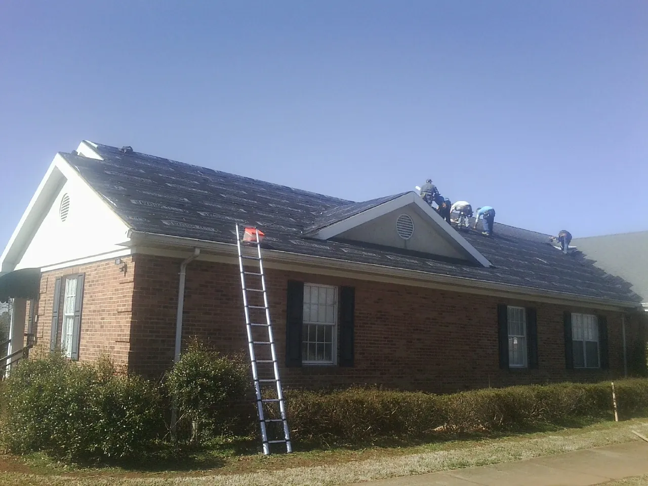 Crew tearing shingles off a brick ranch roof with a ladder set — Emergency Roof Repair work by our crew in Gate City