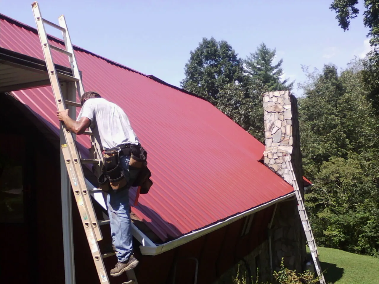 Roofer on a ladder beside a red metal roof and stone chimney — Soffit & Fascia Repair by Roofing Brothers in Gate City